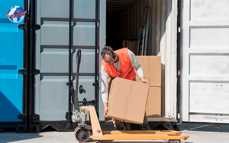A worker loads a large box onto a pallet jack from a shipping container