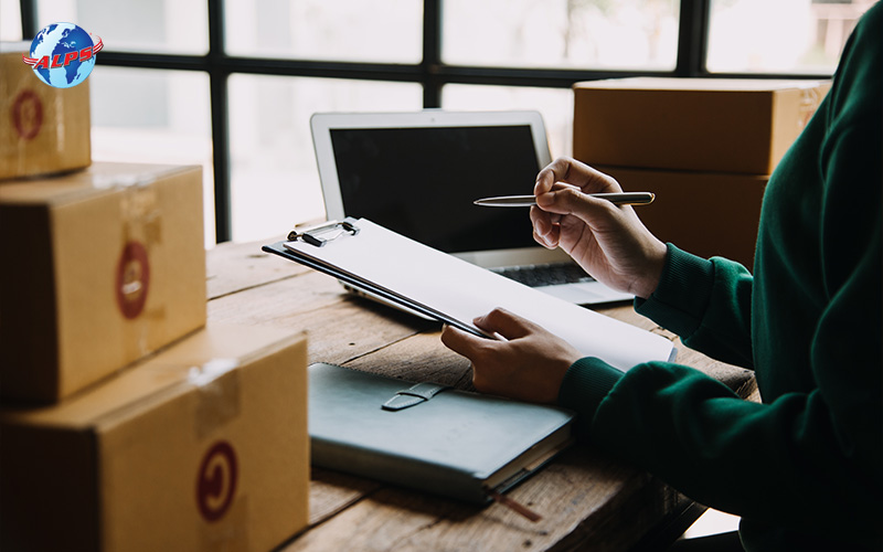 Person writing on a clipboard with packed boxes and a laptop nearby